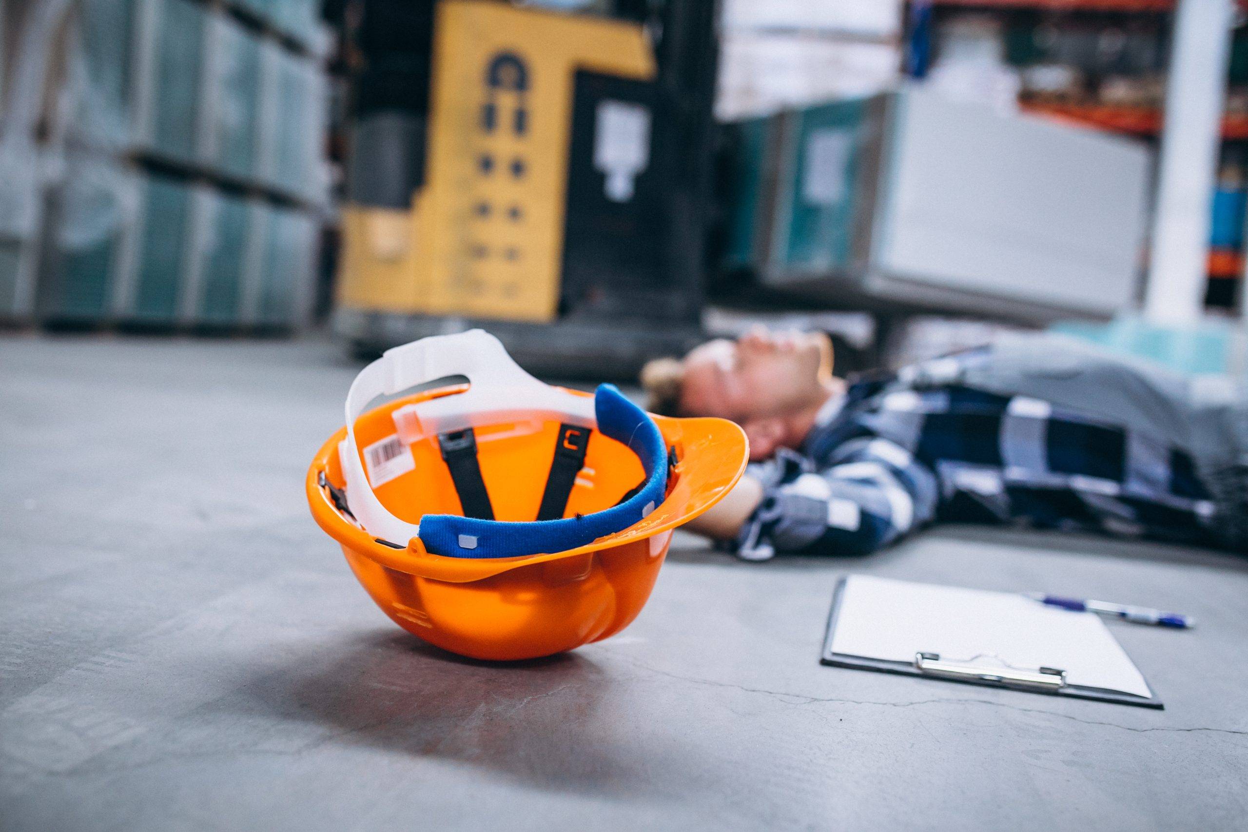 Foto representativa de um acidente de trabalho. Um capacete amarelo, uma prancheta com folhas, uma caneta e mais ao fundo um homem branco com camisa quadriculada estão no chão.